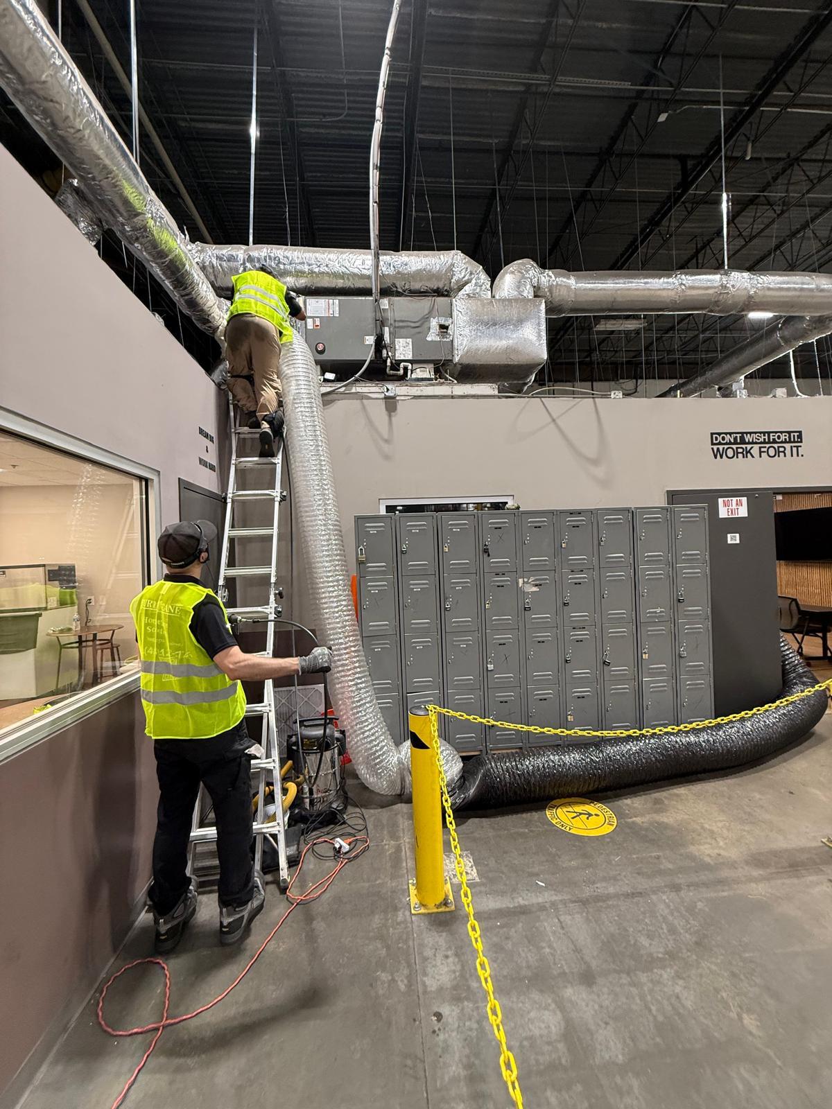Manufacturing floor — two-man crew running negative-air flex from RTU through the break area